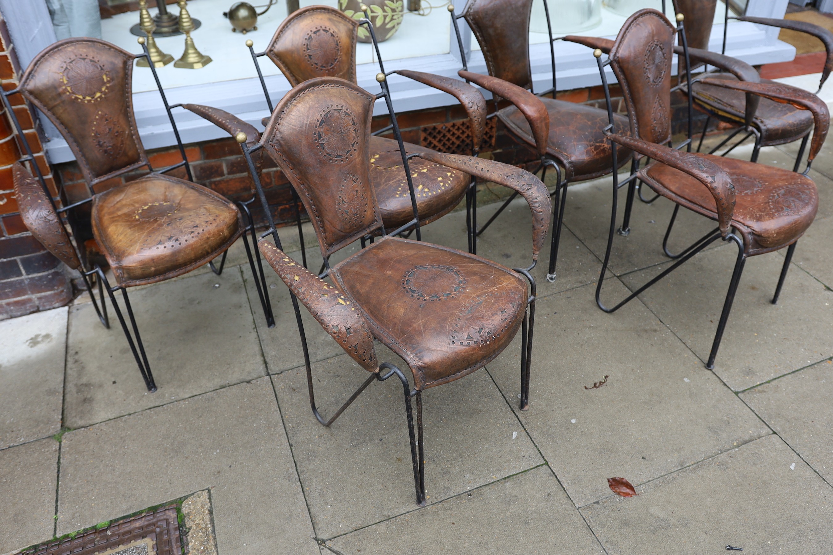 A set of six French wrought iron and brown leather elbow chairs, with brass ball finials, four with tooled and pierced decoration the others plain, height 84cm. width 61cm